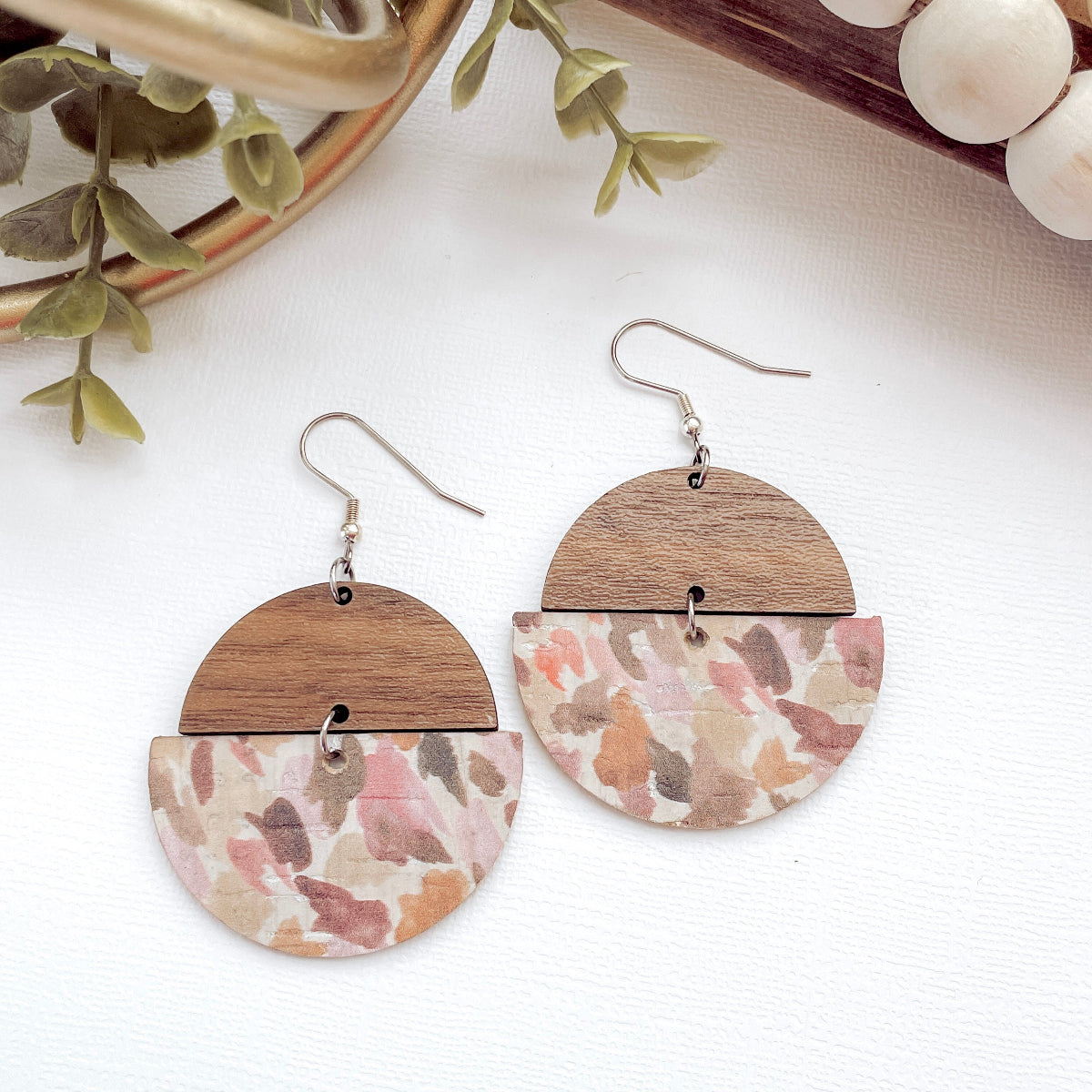 Wooden and floral-patterned earrings on a white surface with greenery and beads in the background.
