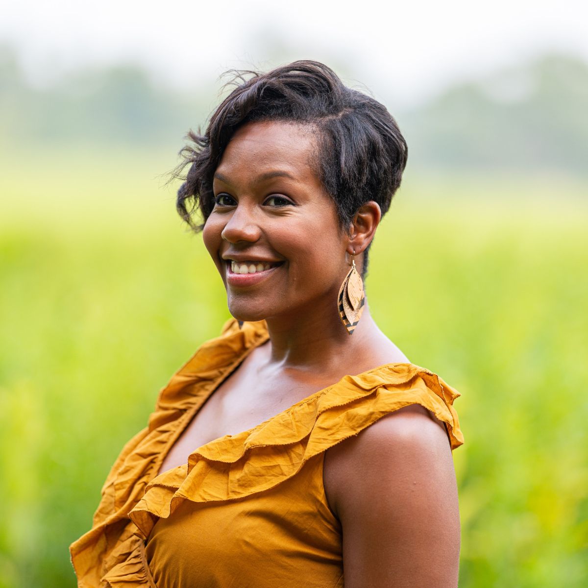 Woman wearing a yellow top with a blurred green background and creek+elm macy cork leaf earrings