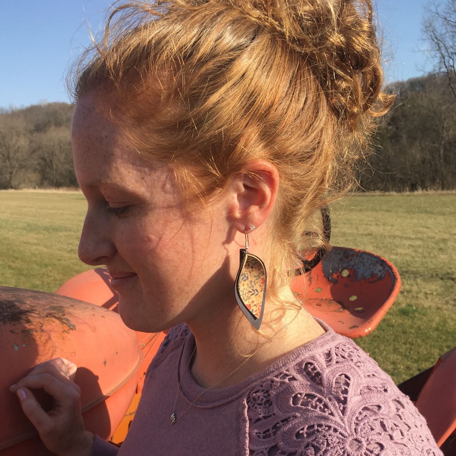 A woman outdoors wearing a pair of large, teardrop-shaped earrings with a black frame and a colorful, textured mosaic center. She is wearing a lavender lace top, with orange farm equipment and a grassy field in the background.