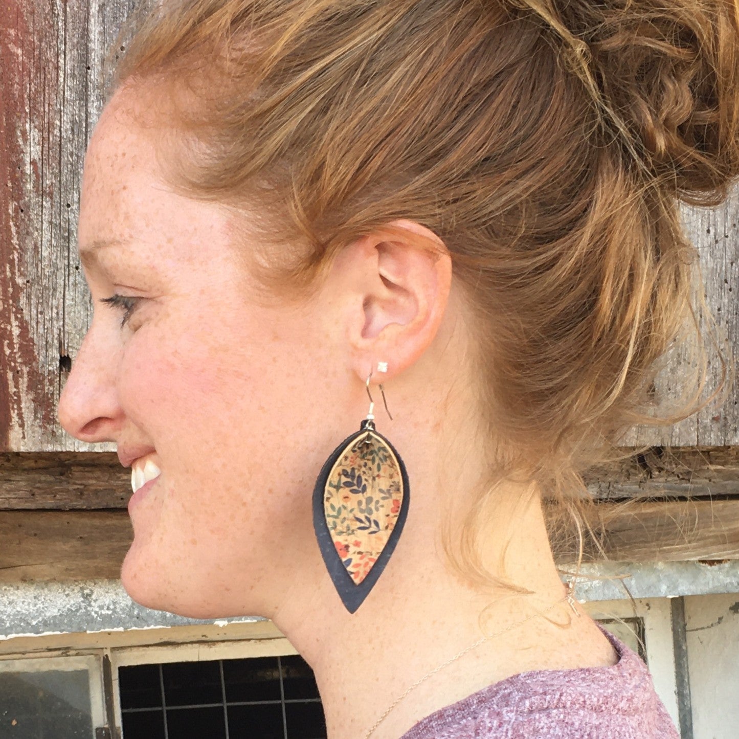 A woman shown in profile wearing a large teardrop-shaped dangle earring with a black border and a colorful patterned center, photographed against a rustic weathered wood background.