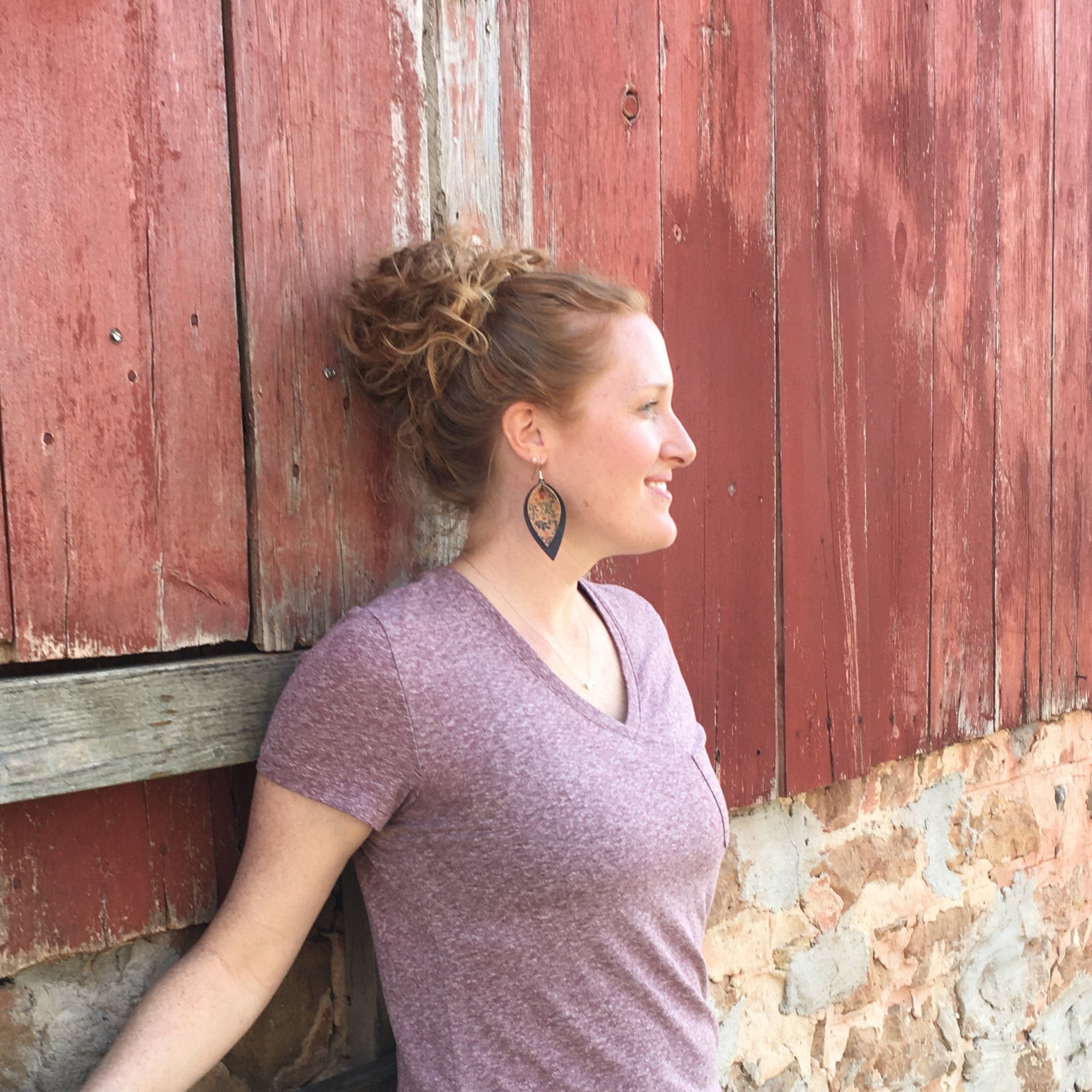 A woman with her hair in a messy bun leans against a weathered red wooden barn wall, wearing a heathered mauve-purple V-neck t-shirt and dark brown teardrop-shaped earrings.