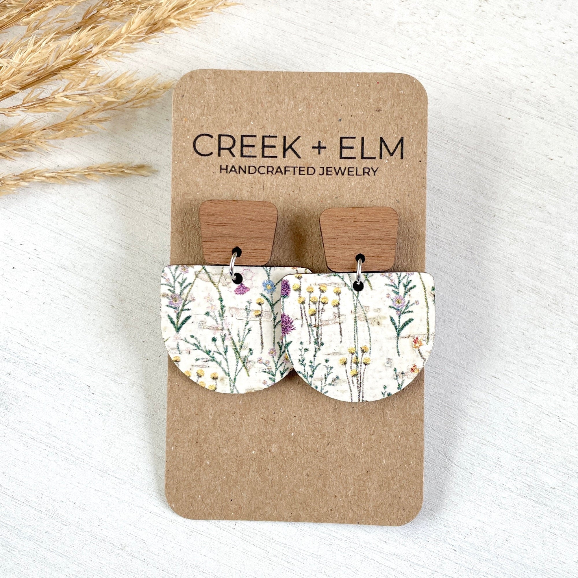 Pair of earrings on a Creek + Elm branded card with dried plants in the background