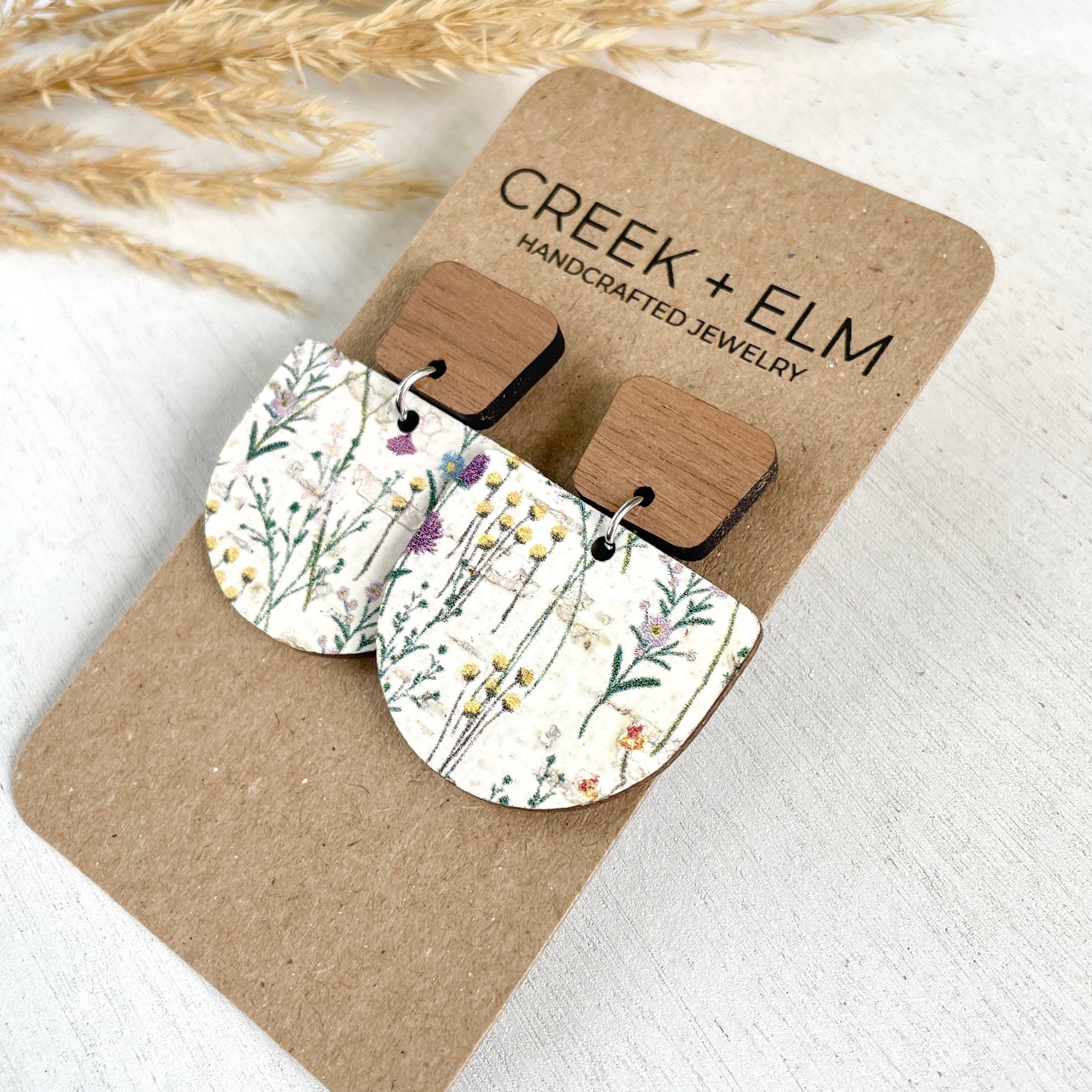 Pair of floral cork and walnut earrings on a 'Creek + Elm' branded card with dried grasses in the background.