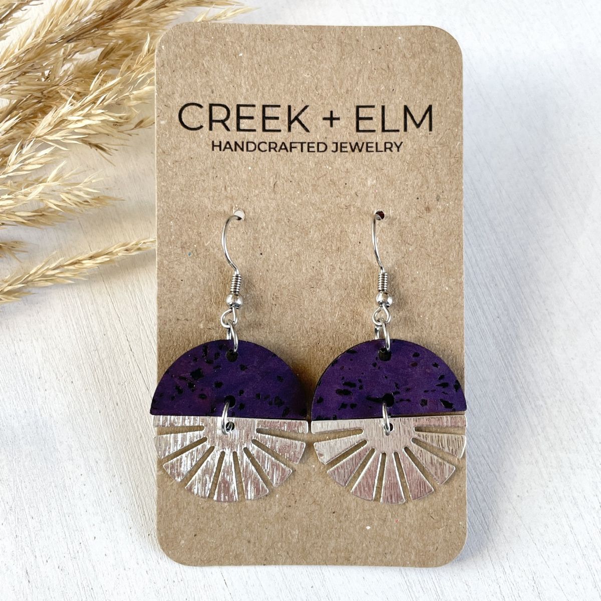 Eggplant-toned earrings with silver starburst accent and soft finish on packaging 