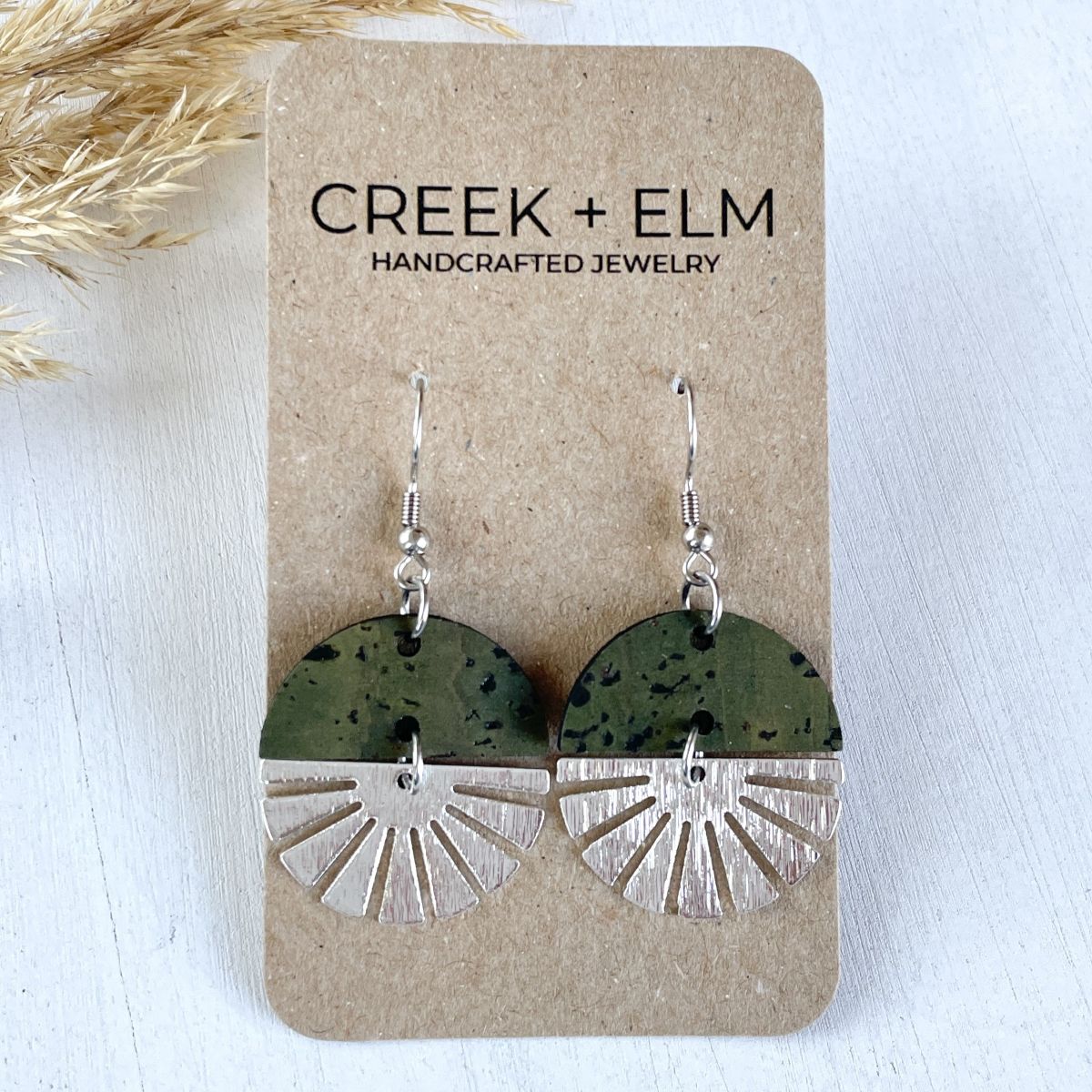 Olive-toned earrings with silver starburst detail and soft natural finish on a packaging