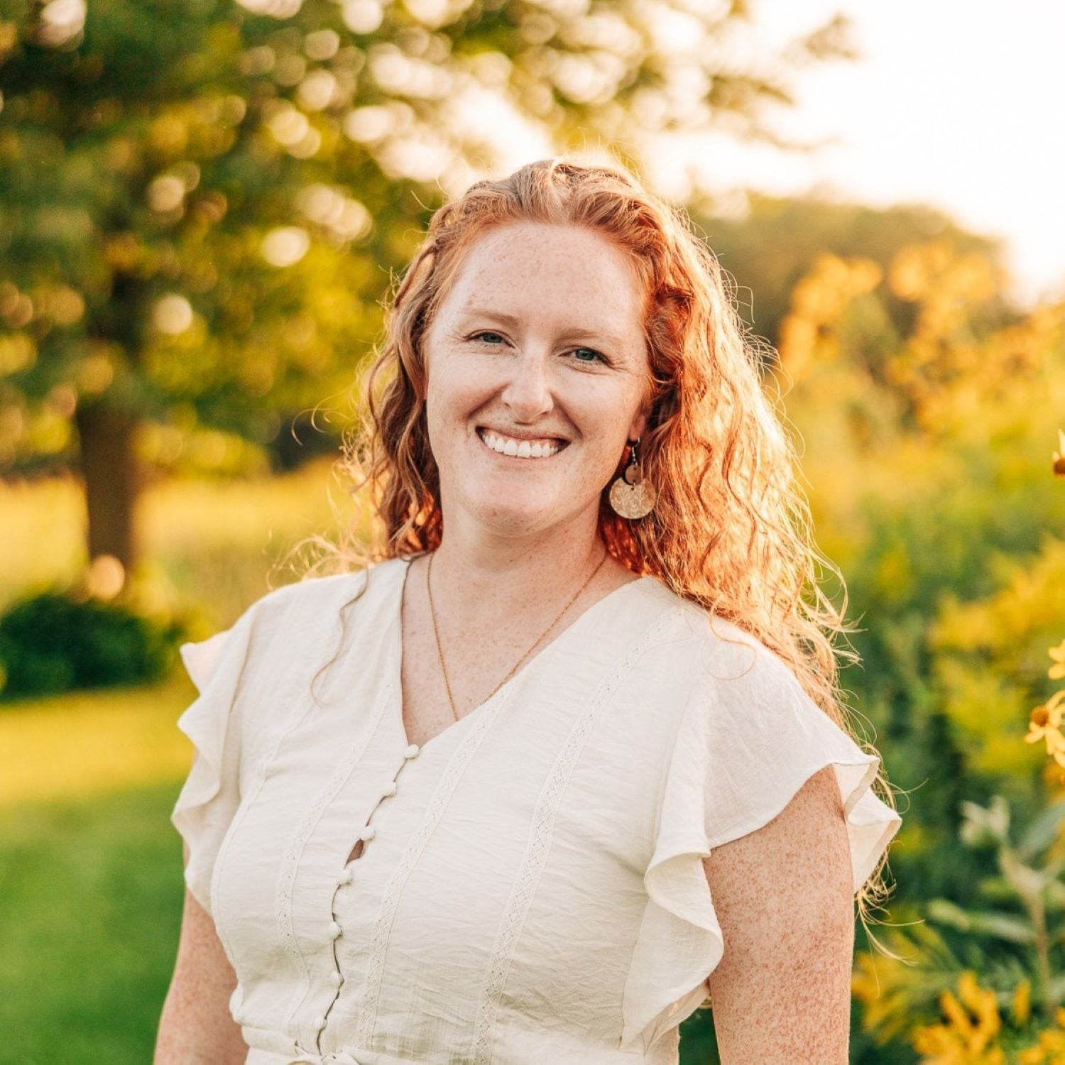 A smiling woman with curly red hair wearing a cream-colored short-sleeve blouse, standing outdoors in a sunlit garden with blurred green trees and foliage in the background.
