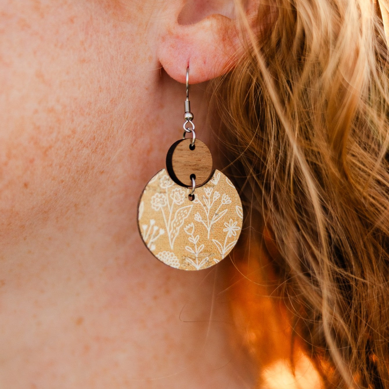 A close-up of a person's ear wearing a dangling earring. The earring features a silver fishhook wire, a small dark wood disc, and a larger circular drop with a beige background and white floral line pattern.
