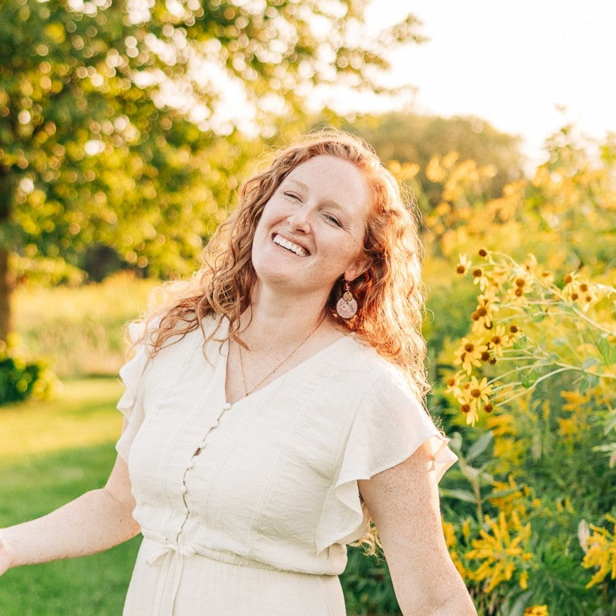 A smiling woman with curly red hair stands outdoors in a sunlit garden wearing a cream-colored short-sleeve dress. Yellow flowers and green foliage fill the background.