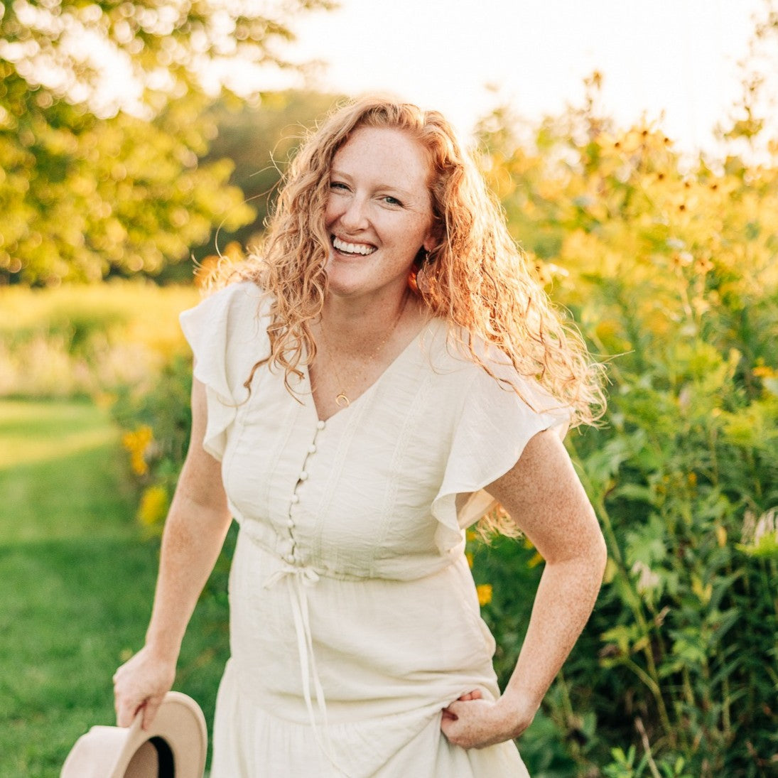 A smiling woman with curly red hair stands outdoors in a sunlit garden wearing a cream-colored dress and holding a light beige hat.