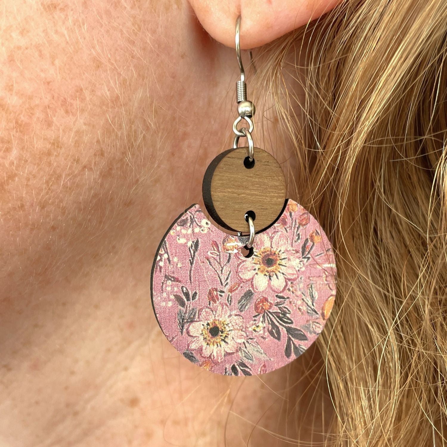 A close-up of a person's ear wearing a dangling earring. The earring has a silver fishhook wire, a round dark wood disc, and a larger circular charm printed with a pink floral pattern.