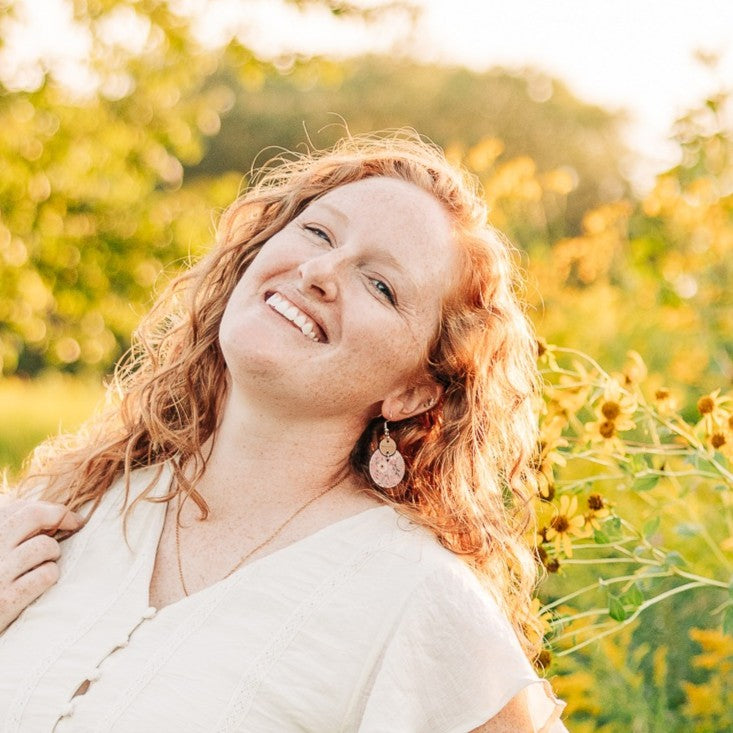A smiling woman with curly red hair wearing a white top and dangling earrings, photographed outdoors in a sunlit field with green and yellow flowers blurred in the background.