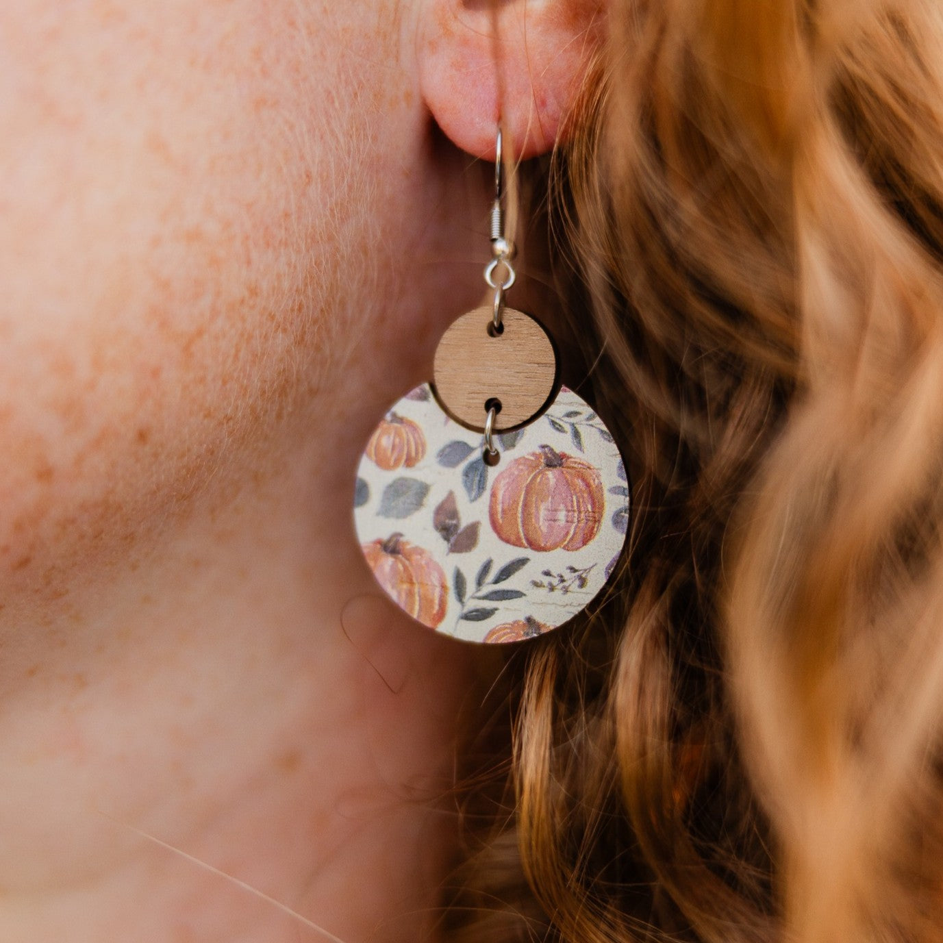 A close-up of a person's ear wearing a dangling earring. The earring has a silver fishhook wire, a small round natural wood disc, and a larger circular charm printed with a fall pumpkin pattern in orange, brown, and muted green on a cream background.