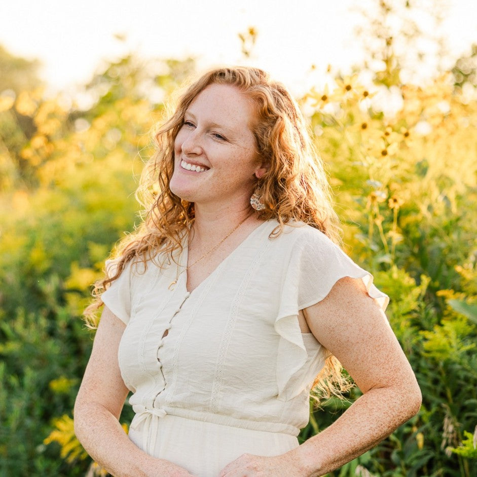 A smiling woman with curly red hair stands outdoors in a sunlit field, wearing a light cream short-sleeve wrap-style dress with ruffled shoulders and a front tie detail.