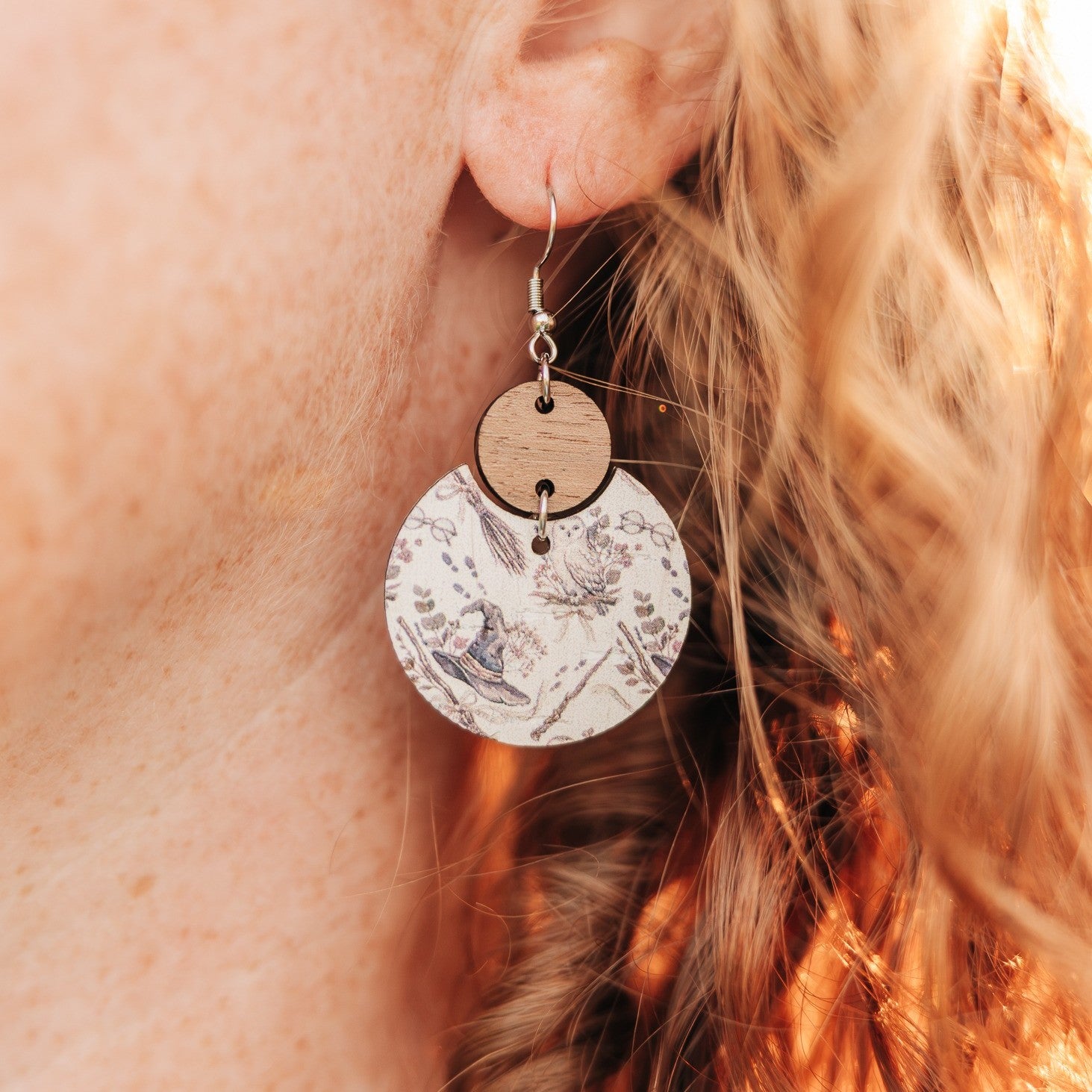 A close-up of a person's ear wearing a dangling earring. The earring has a silver fishhook wire, a small round natural wood disc, and a larger circular charm printed with a vintage-style botanical and witch illustration in muted blue-gray tones on a white background.