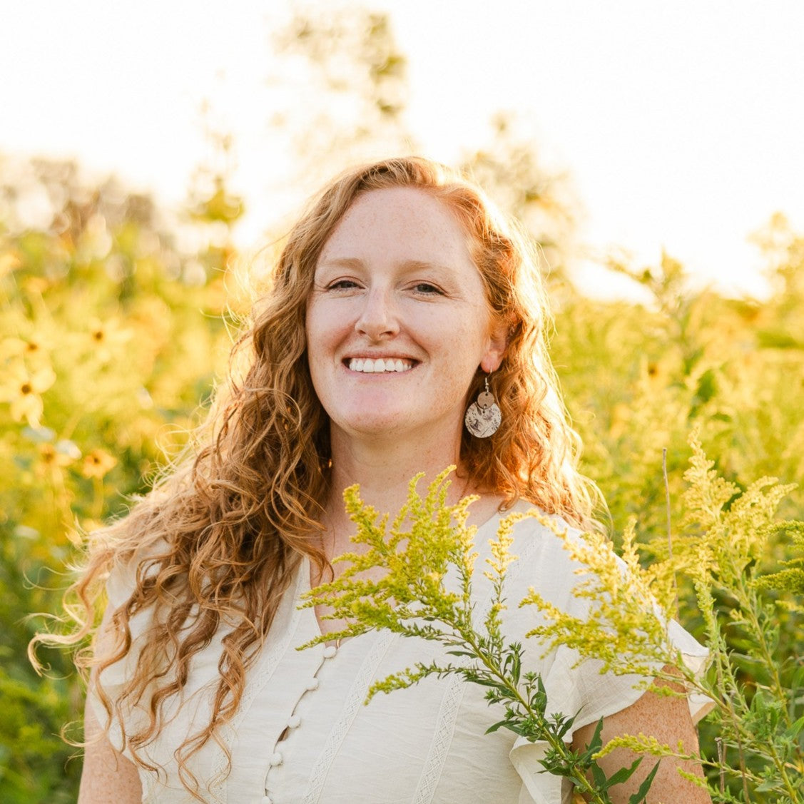 A smiling woman with long curly light brown hair stands outdoors in a sunlit field of yellow wildflowers, wearing a cream short-sleeve top and dangling earrings.