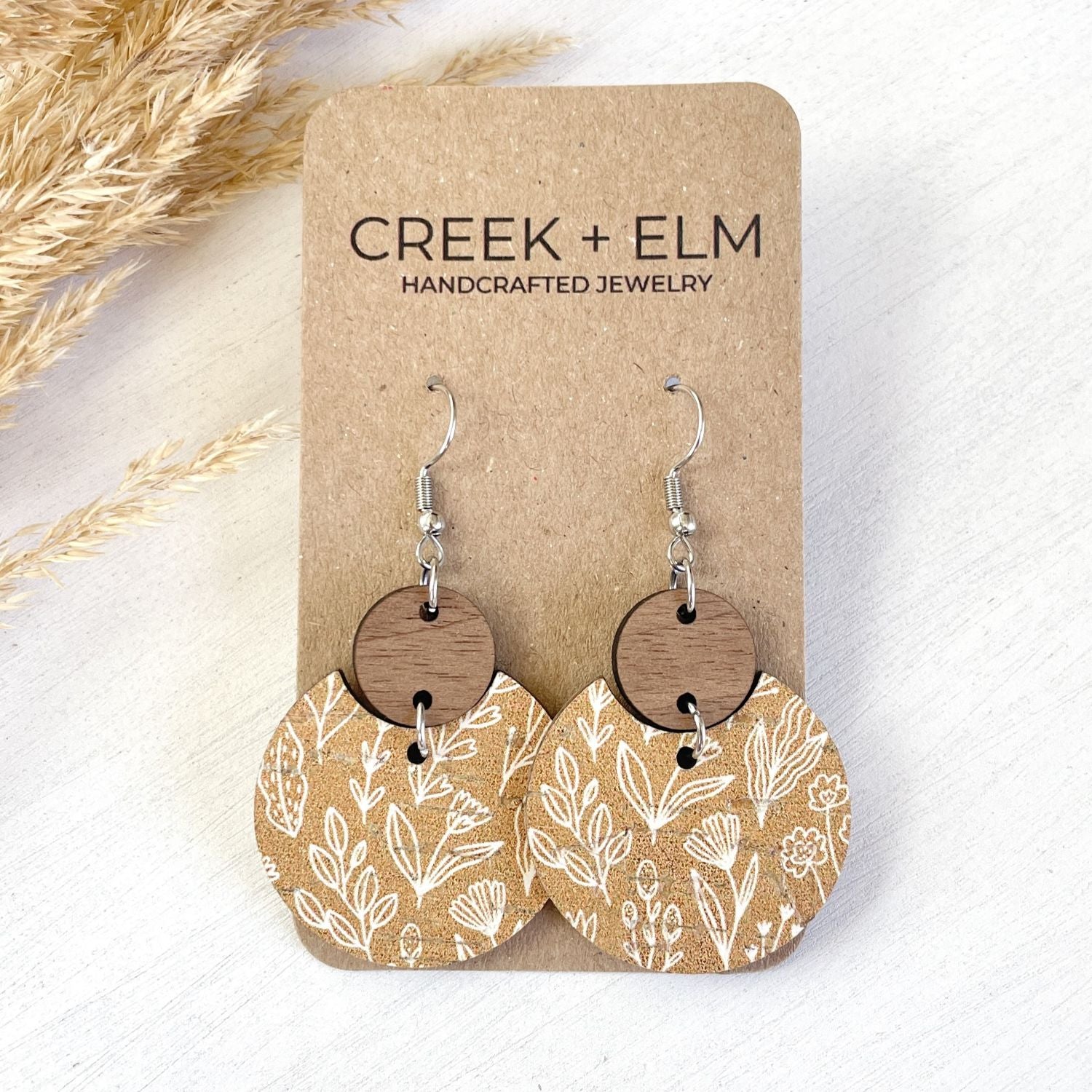 A pair of handmade drop earrings displayed on a kraft paper card. The earrings feature silver-tone fishhook wires, round natural wood discs, and circular tan paper charms printed with a white botanical leaf pattern.