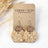 A pair of handmade drop earrings displayed on a kraft paper card. The earrings feature silver-tone fishhook wires, round natural wood discs, and circular tan paper charms printed with a white botanical leaf pattern.