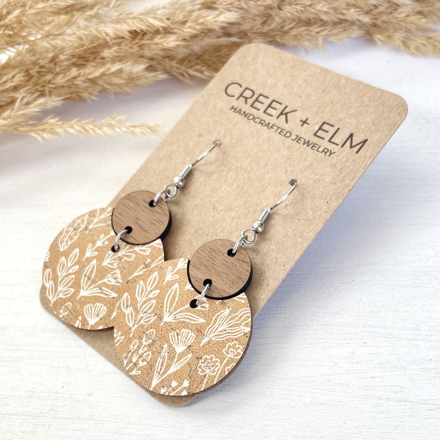 Pair of handmade dangle earrings displayed on a kraft card. The earrings feature circular wooden discs with a natural wood grain top and bottom, connected by a silver-toned wire, with oval wooden charms printed with a white botanical leaf pattern.