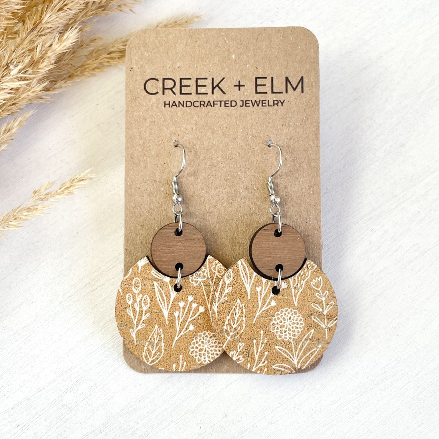 A pair of handmade dangle earrings displayed on a brown kraft card. The earrings feature silver-toned fishhook wires, round natural wood discs, and irregular scalloped pieces with a white botanical line-art pattern on a tan background.