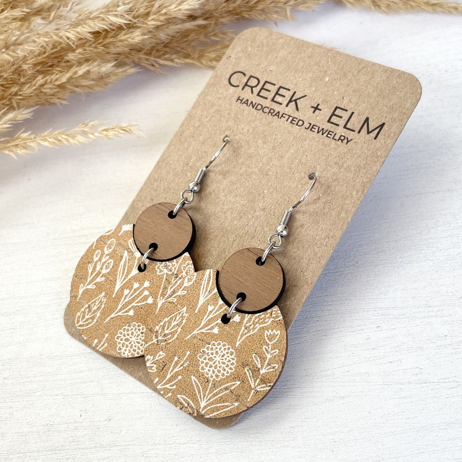 Wooden dangle earrings with a floral pattern on the lower half and a round wooden disc on the upper half, displayed on a kraft paper card labeled CREEK + ELM HANDCRAFTED JEWELRY.
