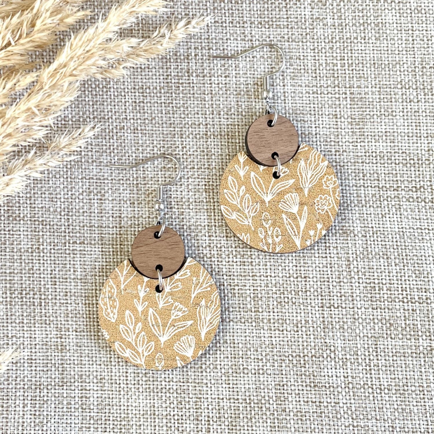 A pair of bohemian-style dangle earrings with circular wooden discs printed with a white botanical pattern on a tan background, hanging from silver fishhook wires on a textured beige linen fabric backdrop with dried grass in the corner.