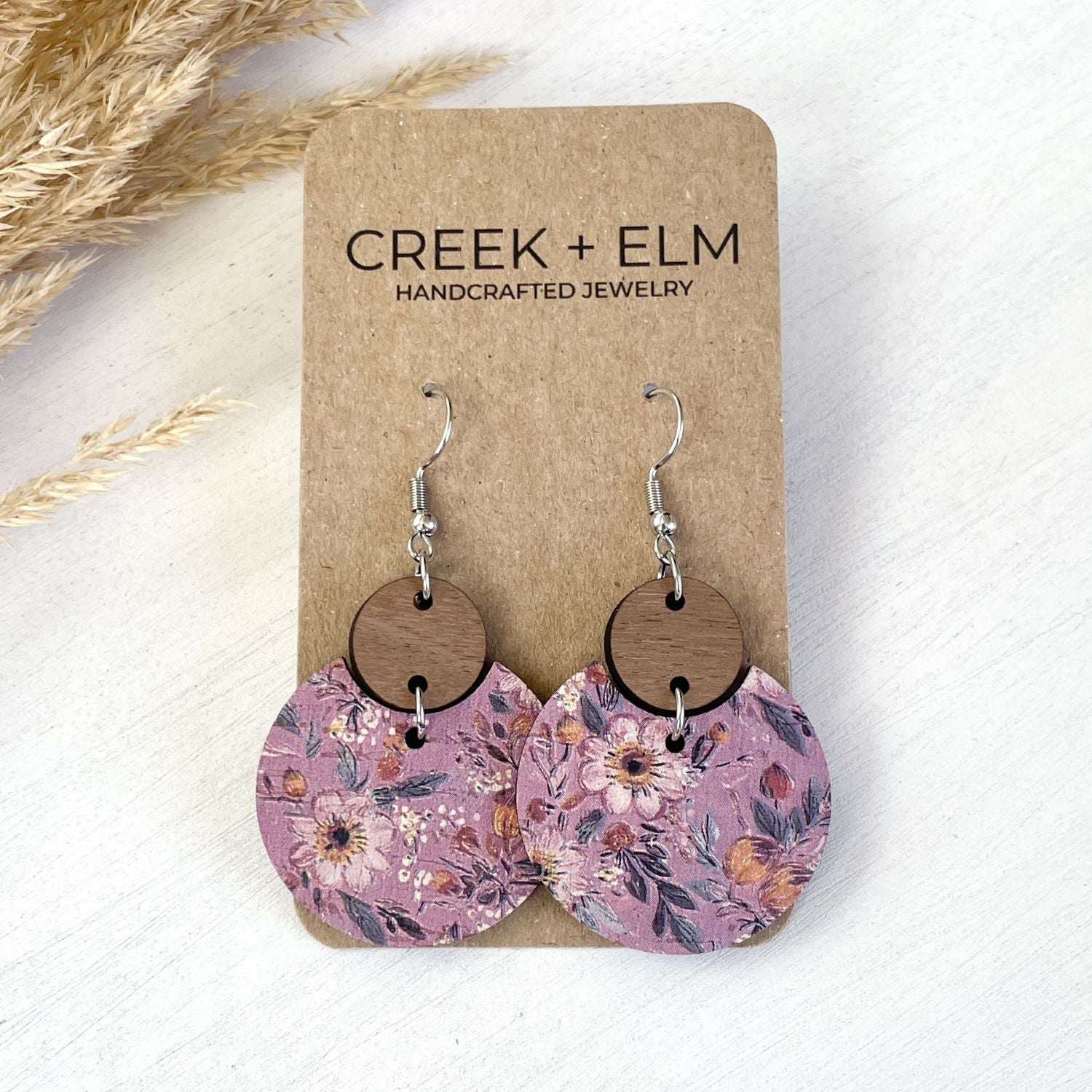 A pair of handmade dangle earrings displayed on a brown kraft card. The earrings feature round floral-patterned wooden discs in dusty pink and purple tones, with silver fishhook wires.