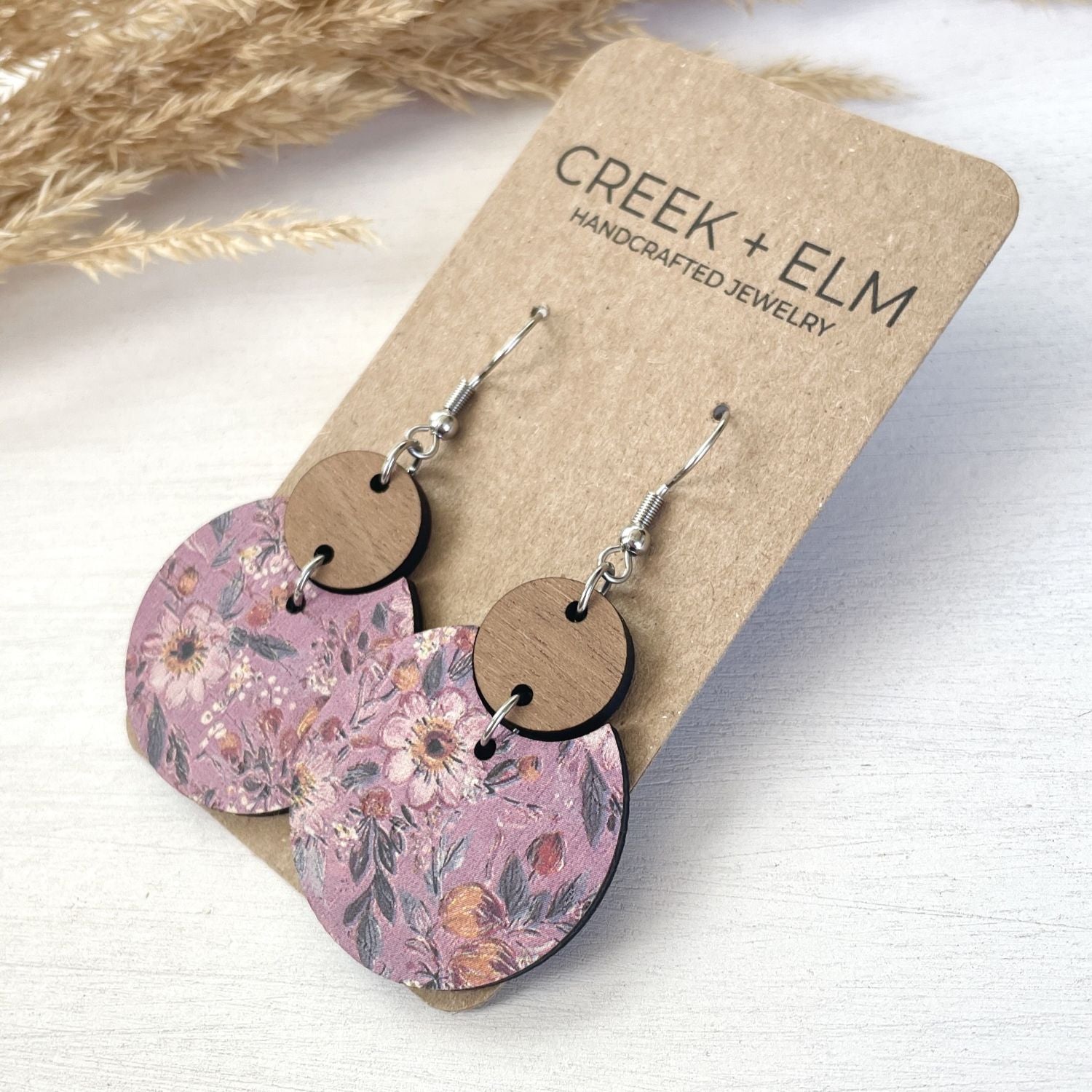 Pair of handmade dangle earrings with floral-patterned circular drops and natural wood discs, displayed on a kraft card labeled CREEK + ELM, on a light wood surface with dried grass in the background.