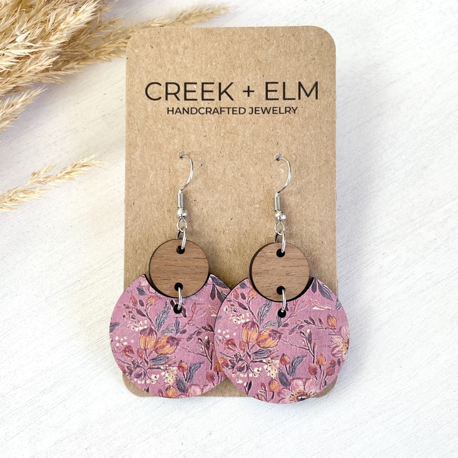 A pair of handmade dangle earrings displayed on a brown kraft card. The earrings feature a dusty pink floral patterned disc hanging from a round natural wood disc and a silver fishhook wire.