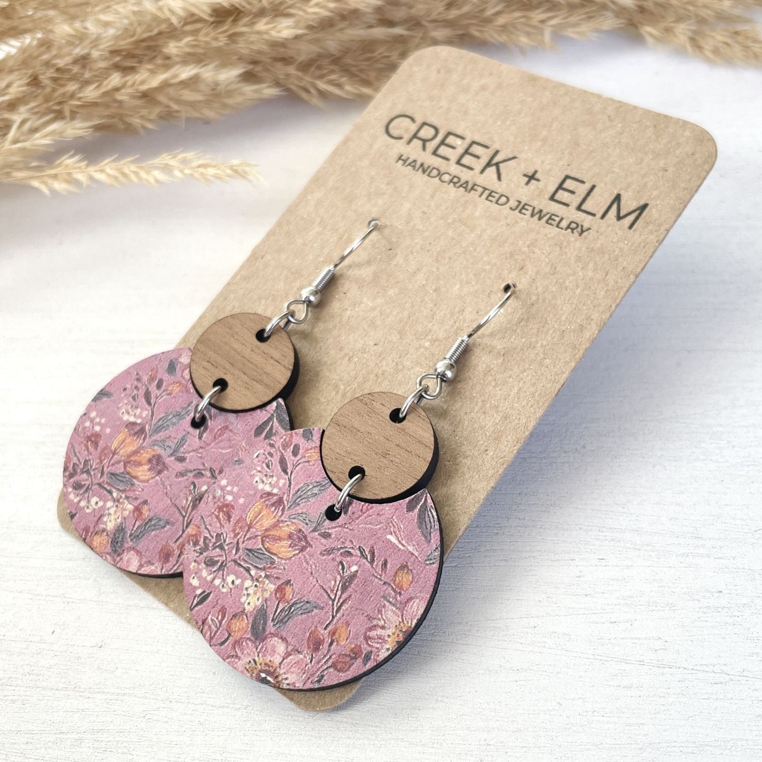 A pair of handmade dangle earrings by Creek + Elm, featuring dusty pink floral patterned wooden discs and round natural wood discs connected by silver wire hooks, displayed on a kraft paper backing card.