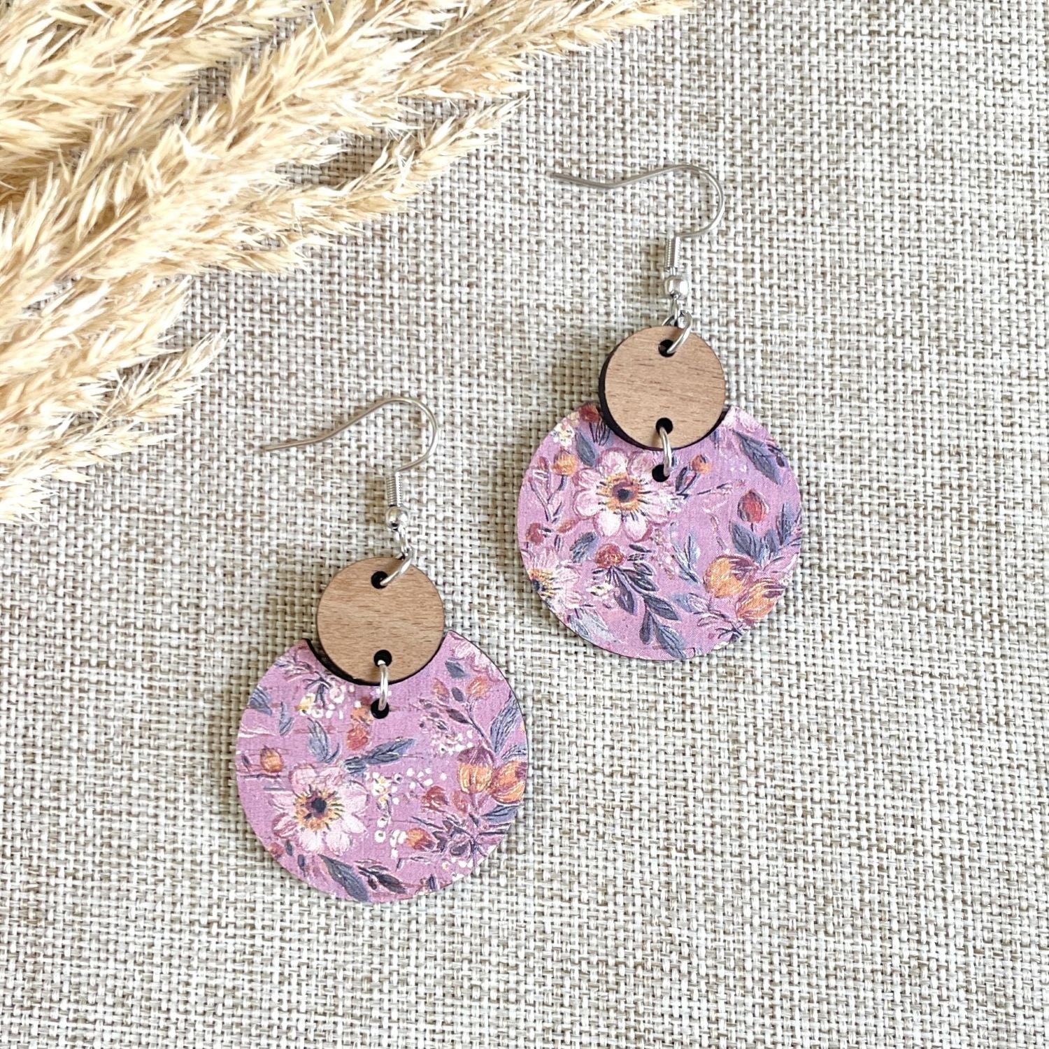A pair of dangling earrings with circular floral prints in pink, purple, and orange tones, hanging from silver fishhook wires with round wooden discs above them, displayed on a textured beige fabric background with dried grass in the top left corner.