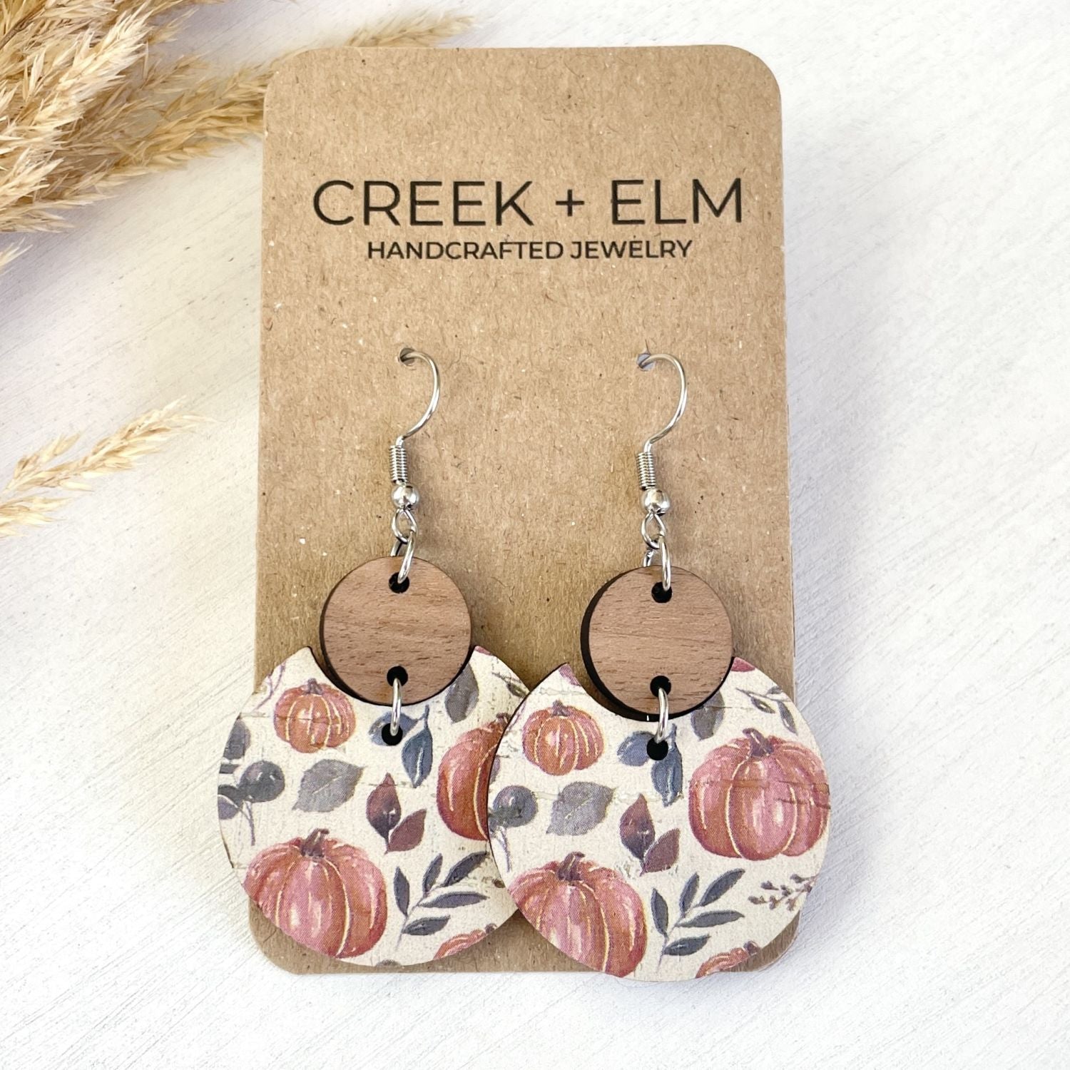 A pair of handmade dangle earrings displayed on a brown kraft card labeled 'CREEK + ELM HANDCRAFTED JEWELRY.' Each earring has a silver fishhook wire, a round natural wood disc, and a larger irregular lower disc printed with a fall harvest pattern of pink pumpkins, muted leaves, and branches on a cream background.