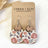 A pair of handmade dangle earrings displayed on a brown kraft card labeled 'CREEK + ELM HANDCRAFTED JEWELRY.' Each earring has a silver fishhook wire, a round natural wood disc, and a larger irregular lower disc printed with a fall harvest pattern of pink pumpkins, muted leaves, and branches on a cream background.