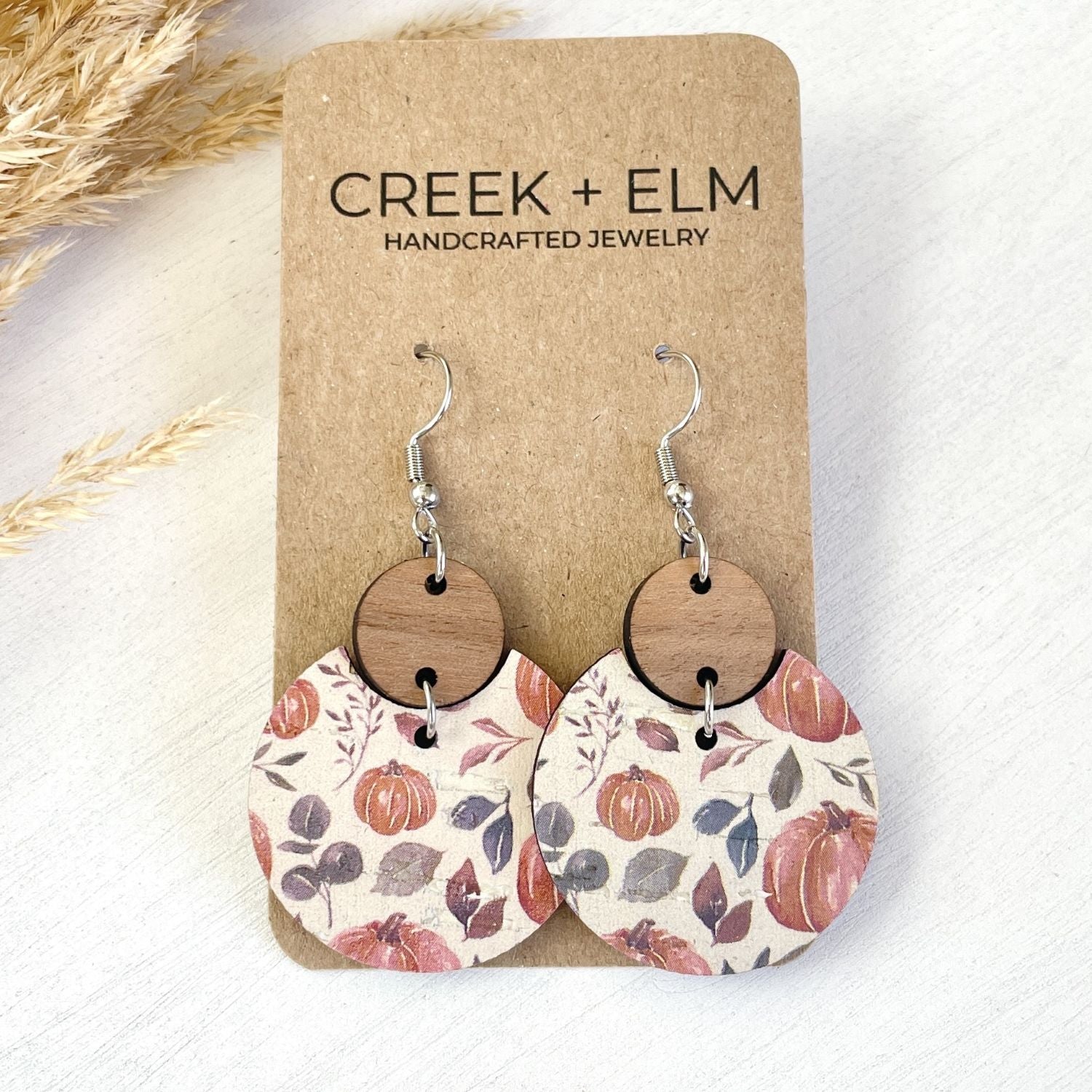 A pair of handmade dangle earrings displayed on a brown kraft card labeled 'CREEK + ELM HANDCRAFTED JEWELRY.' The earrings have silver-tone hooks, round natural wood discs, and irregular lower halves with a fall-themed floral print featuring pumpkins, leaves, and branches in muted orange, mauve, gray, and cream tones.