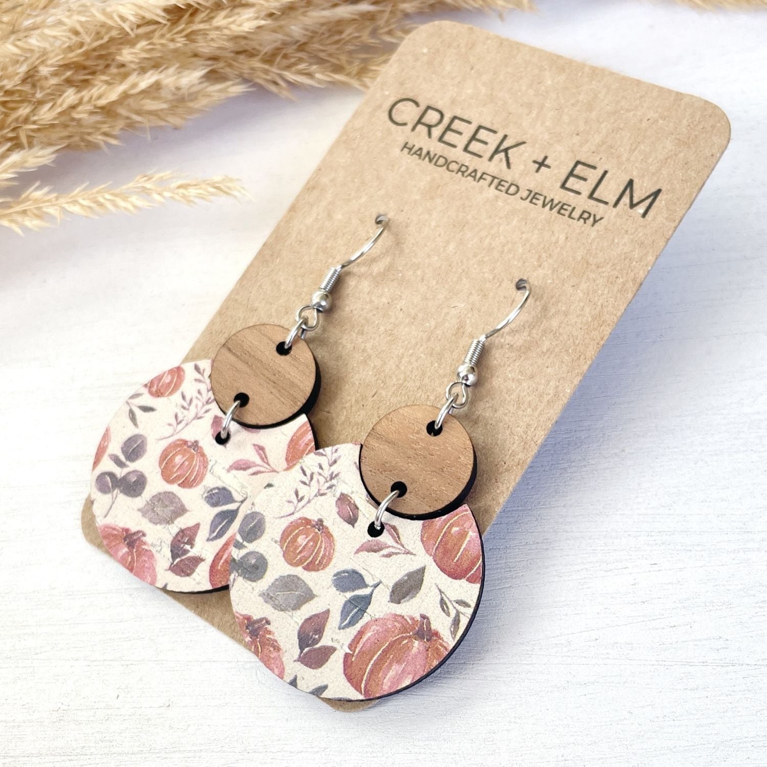 A pair of handmade dangle earrings displayed on a kraft paper card. The earrings feature round natural wood discs attached to silver fishhook wires, with larger scalloped fan-shaped drops printed with a fall harvest pattern of pumpkins, leaves, and acorns in muted red, brown, and gray tones.