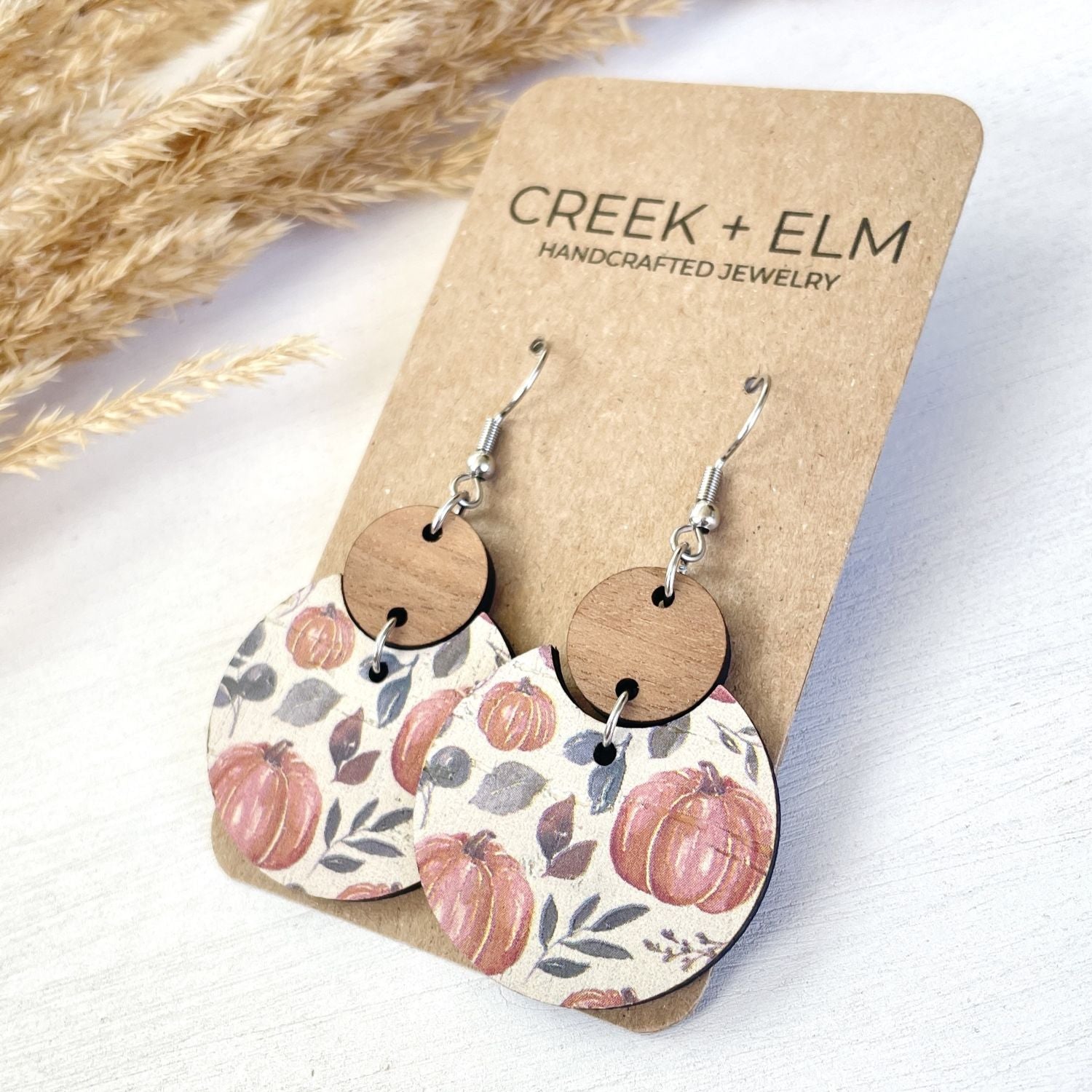 A pair of handmade dangle earrings displayed on a kraft card. The earrings feature silver-toned fishhook wires, round natural wood discs, and semi-circular wooden pendants printed with a fall harvest pattern of pumpkins and leaves on a cream background.