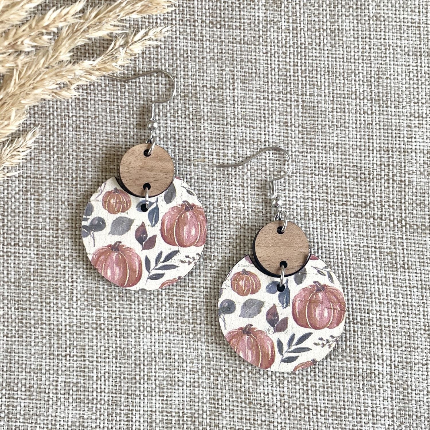 A pair of handmade dangle earrings with round wooden discs and silver fishhook wires, featuring a printed autumn pattern of pumpkins and leaves on a textured beige fabric background.