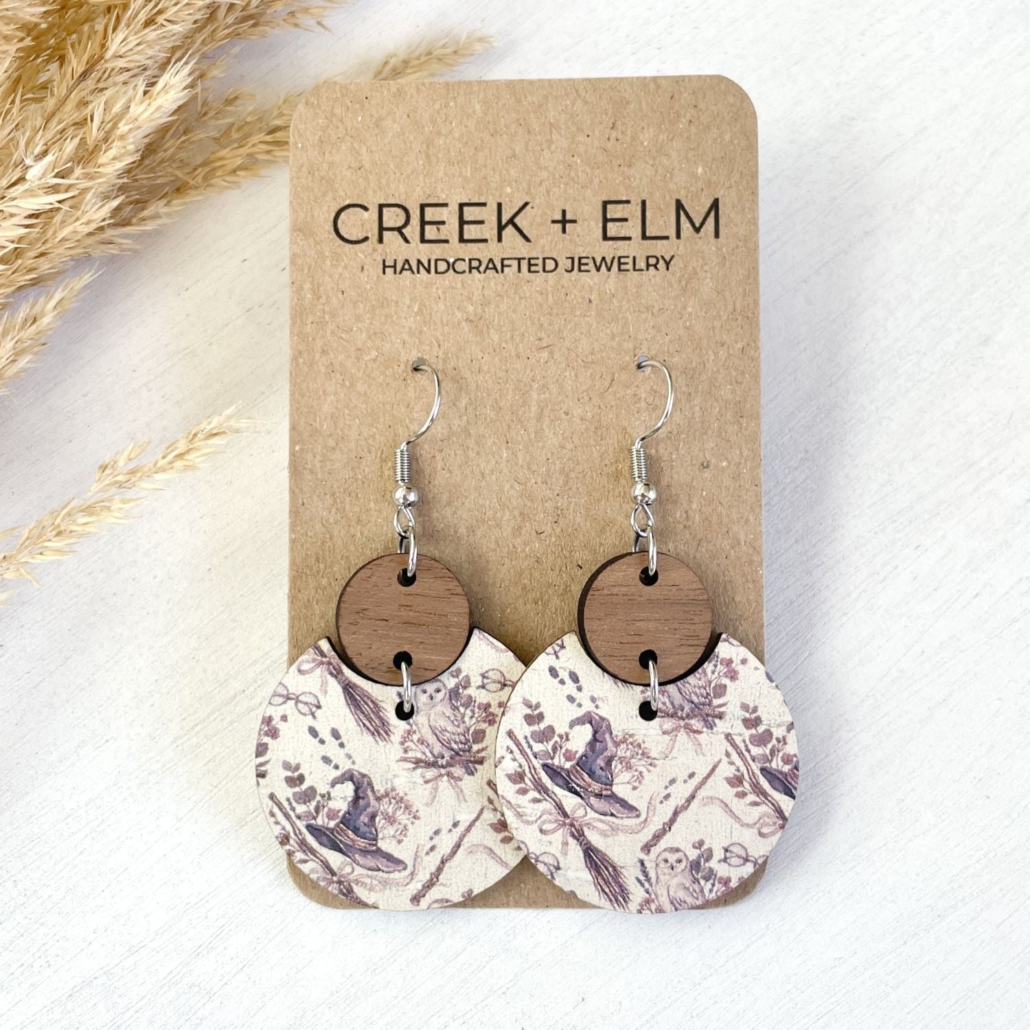 A pair of handmade dangle earrings displayed on a kraft card. The earrings feature circular wooden discs in a medium brown tone, hanging from silver-toned French hook ear wires, with oval wooden charms printed in a muted purple and cream toile-style pattern.