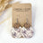 A pair of handmade dangle earrings displayed on a kraft card. The earrings feature circular wooden discs in a medium brown tone, hanging from silver-toned French hook ear wires, with oval wooden charms printed in a muted purple and cream toile-style pattern.