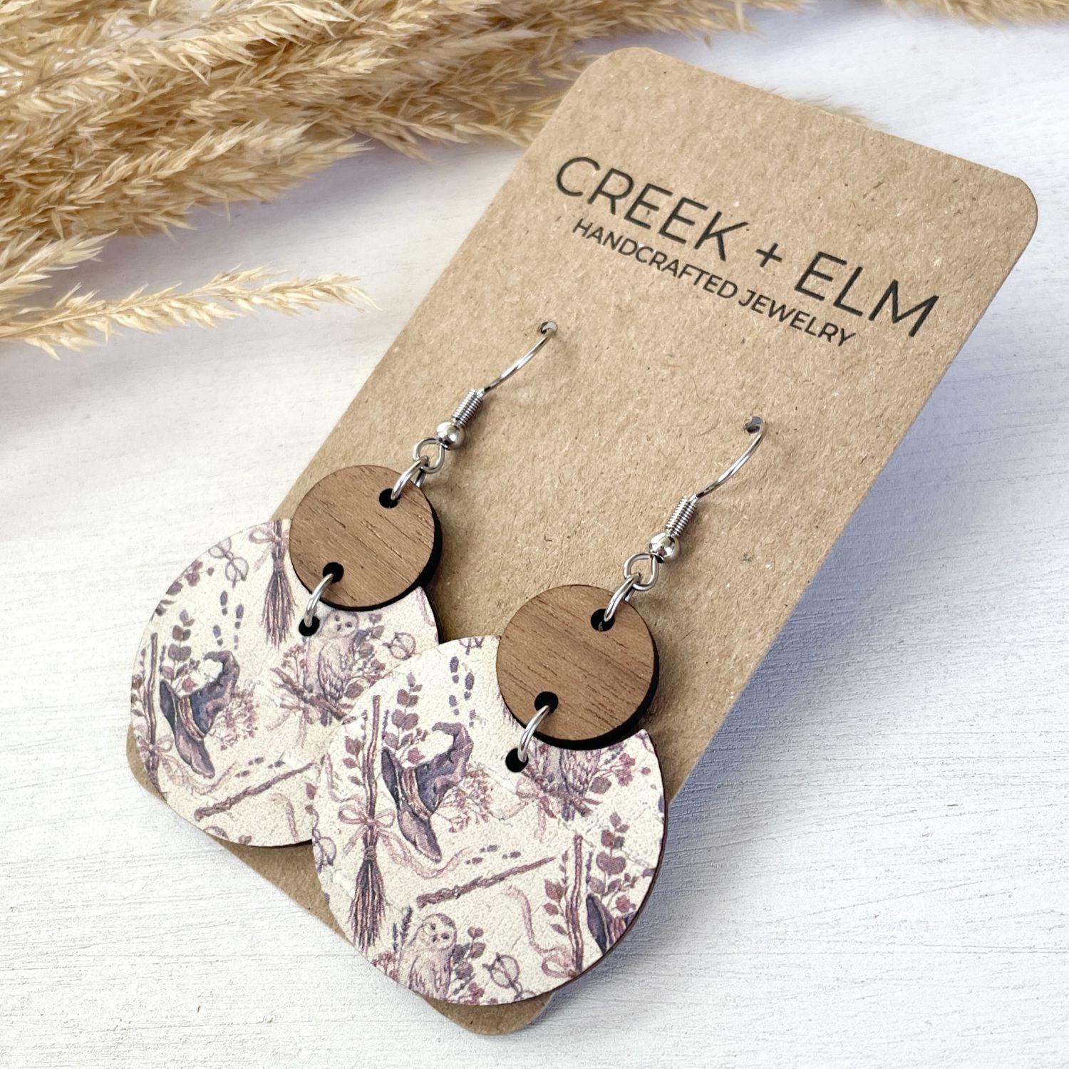 Pair of handmade dangle earrings displayed on a kraft paper backing card. The earrings feature oval wooden discs connected to circular paper discs with a muted floral toile pattern in dusty purple and beige.