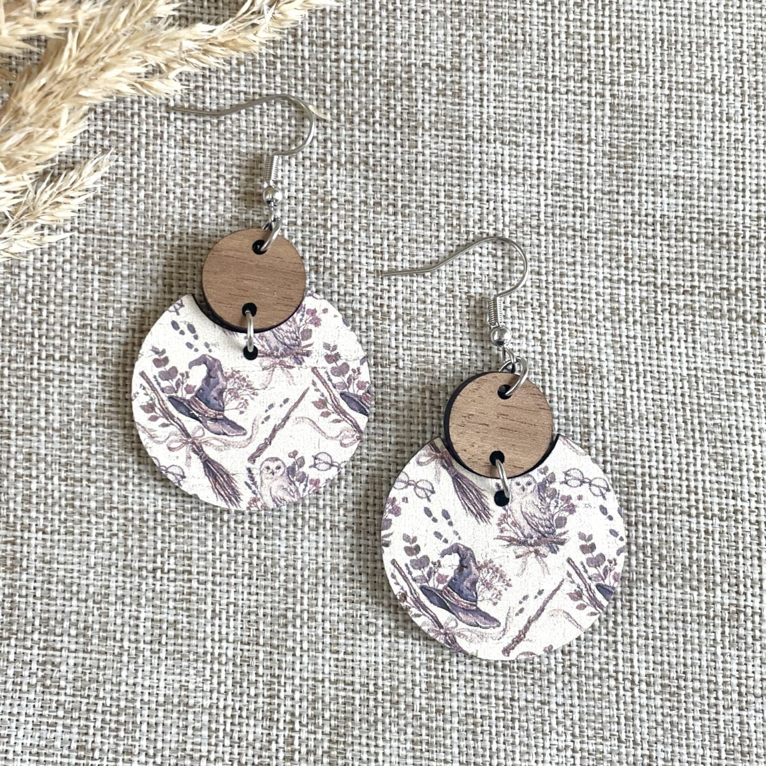 A pair of dangle earrings with round white paper discs printed in a purple botanical toile pattern, attached to round brown wooden discs and silver fishhook wires, displayed on a textured beige linen background.