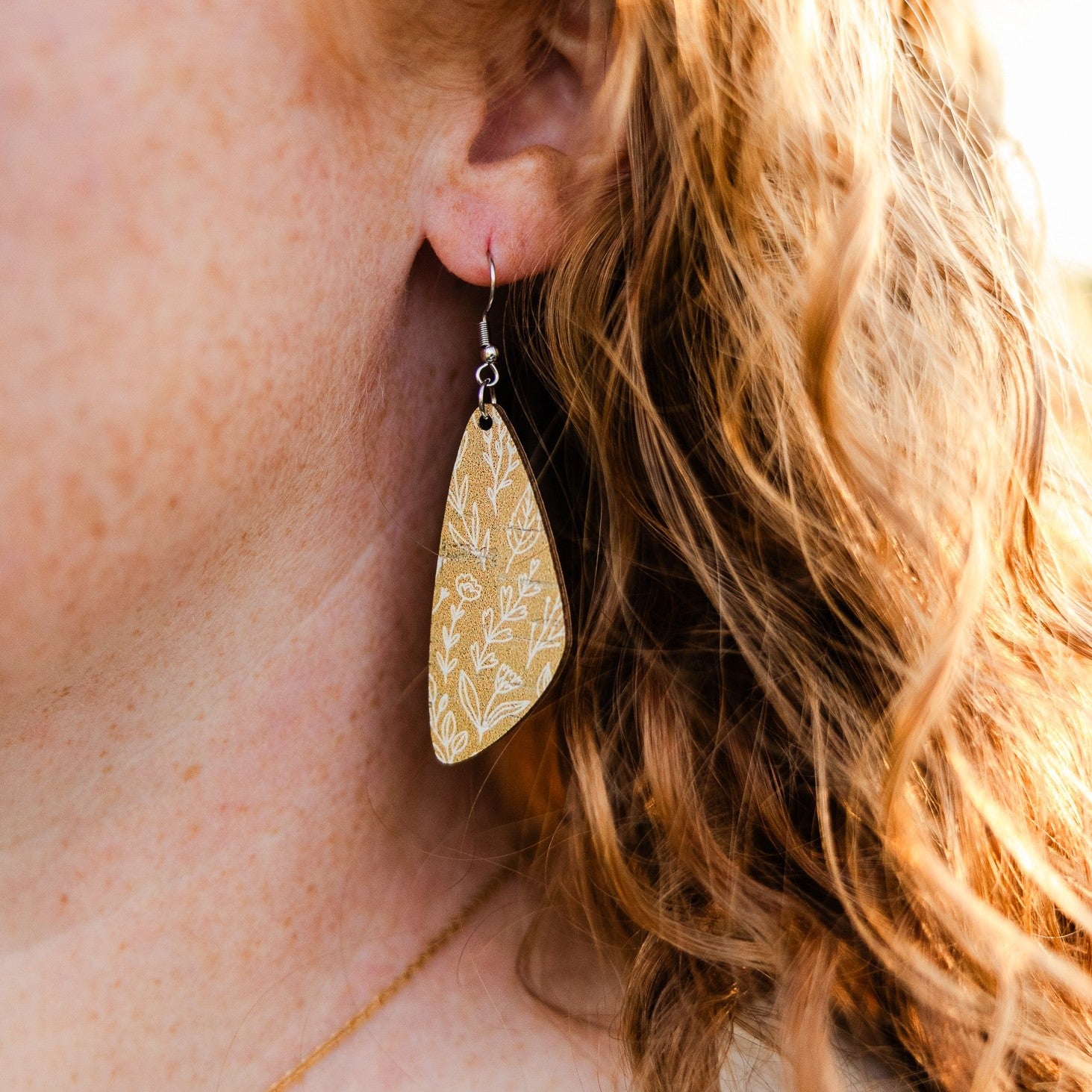 A close-up of a person's ear wearing a teardrop-shaped wooden dangle earring with a white floral pattern on a light wood background.