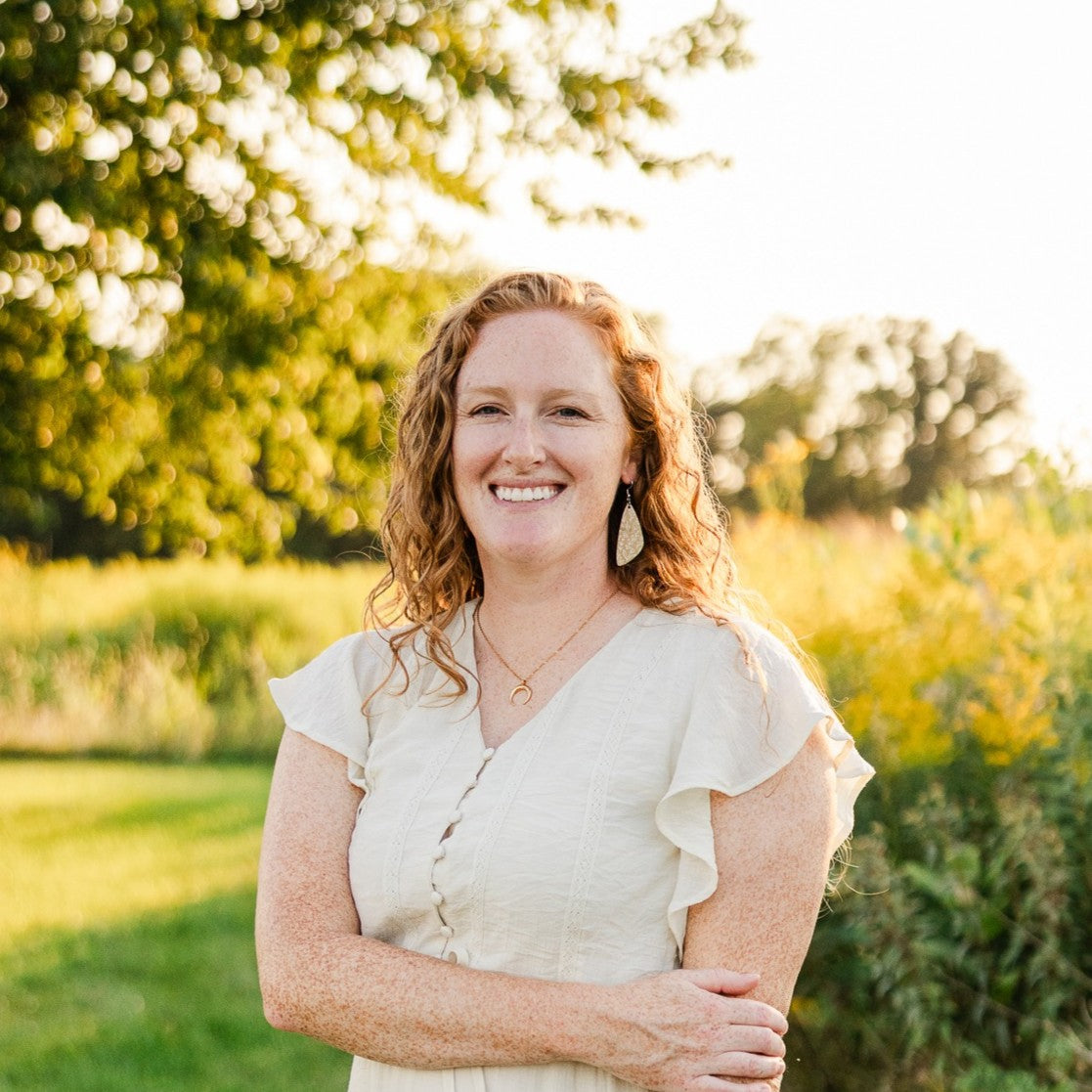 A smiling woman with curly red hair stands outdoors in a sunlit field, wearing a light cream ruffled blouse and gold jewelry.