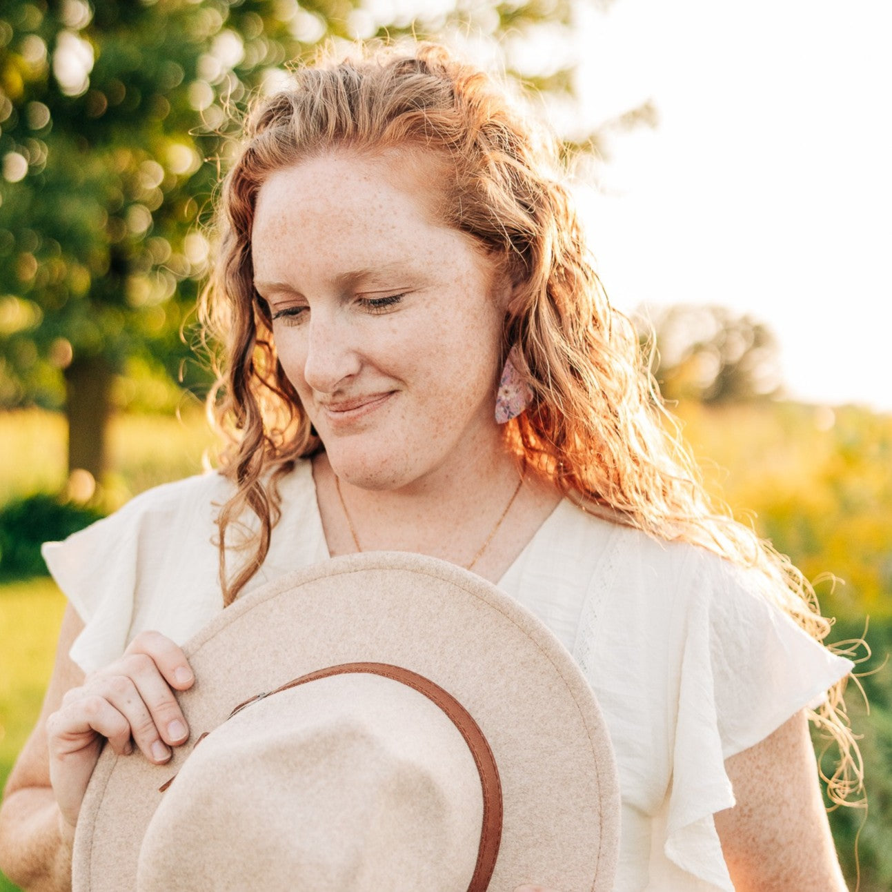 A woman outdoors in warm sunlight wearing a white ruffled blouse and holding a beige wide-brim hat with a brown band. Trees and a sunlit field are blurred in the background.
