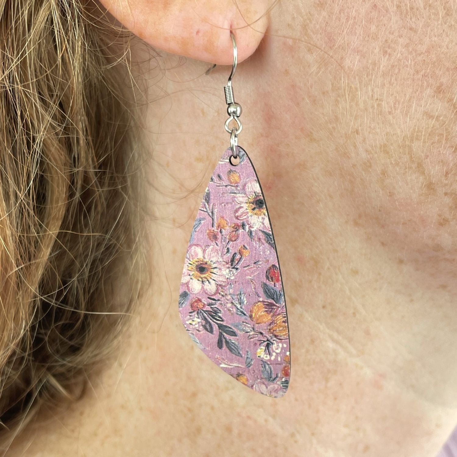 A close-up of a person wearing a teardrop-shaped dangle earring with a pink floral pattern and silver fishhook ear wire.