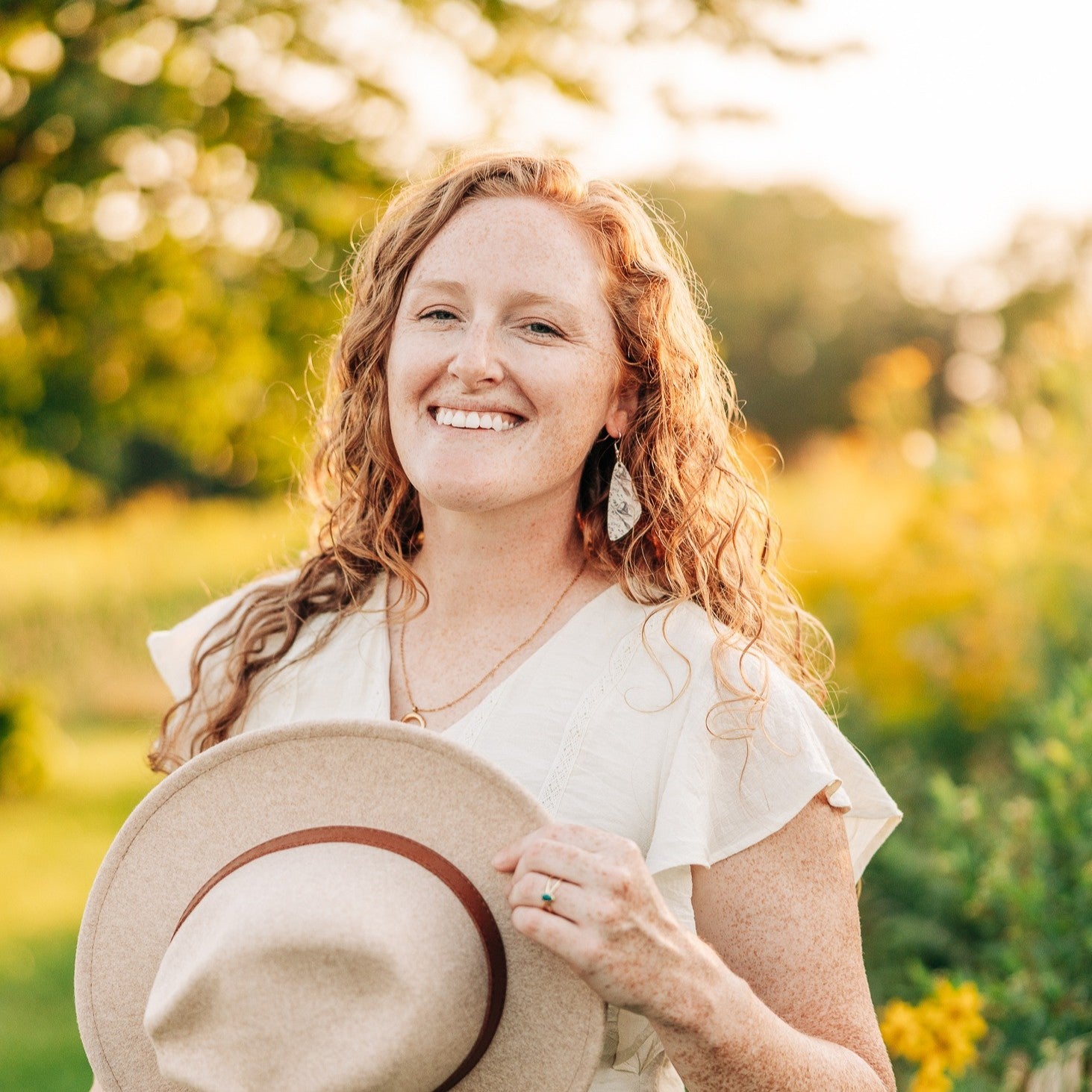 A smiling woman with curly red hair stands outdoors in a sunlit garden, wearing a white ruffled top and holding a light beige wide-brim hat with a brown band.