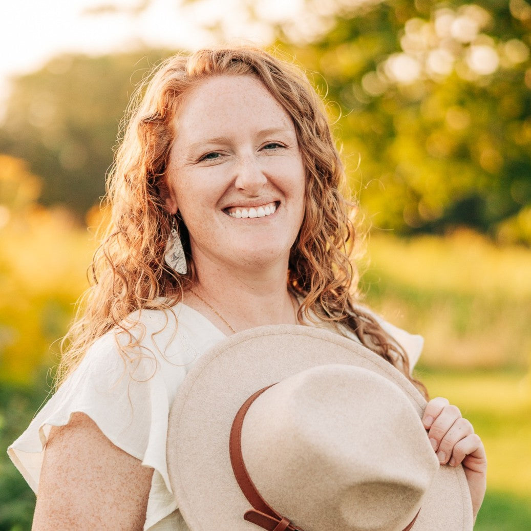 A smiling woman with curly red hair holds a light beige wide-brim hat with a brown band in an outdoor setting with a warm, sunlit bokeh background.