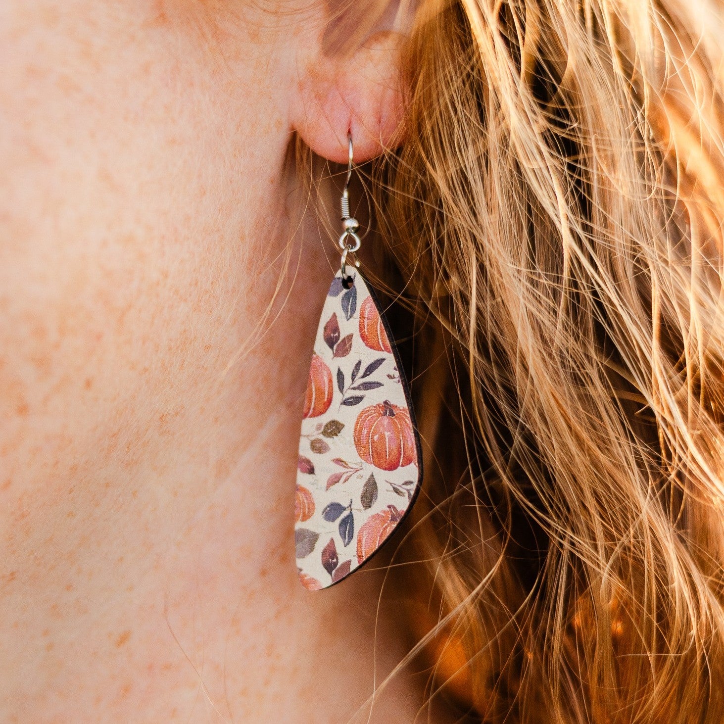 A close-up of a person wearing a teardrop-shaped dangle earring with a fall pumpkin pattern. The earring features orange pumpkins, brown leaves, and muted blue-gray accents on a cream background, suspended from a silver fishhook ear wire.