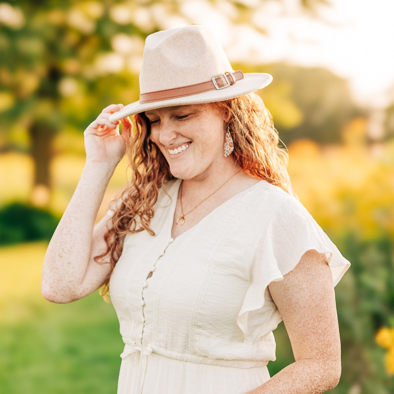 A woman outdoors wearing a light beige wide-brim fedora hat with a brown band and buckle, a cream short-sleeve dress, and gold jewelry, smiling while adjusting the brim of the hat.
