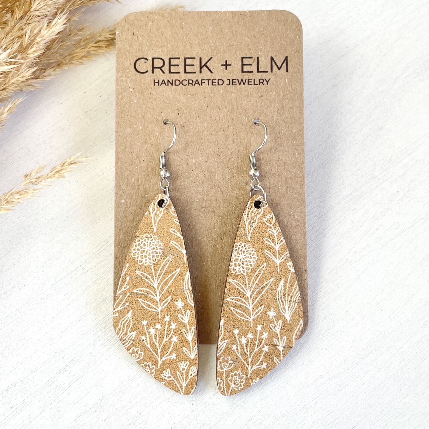 A pair of teardrop-shaped dangle earrings with a white botanical pattern on a tan background, displayed on a brown kraft card labeled 'CREEK + ELM HANDCRAFTED JEWELRY'.