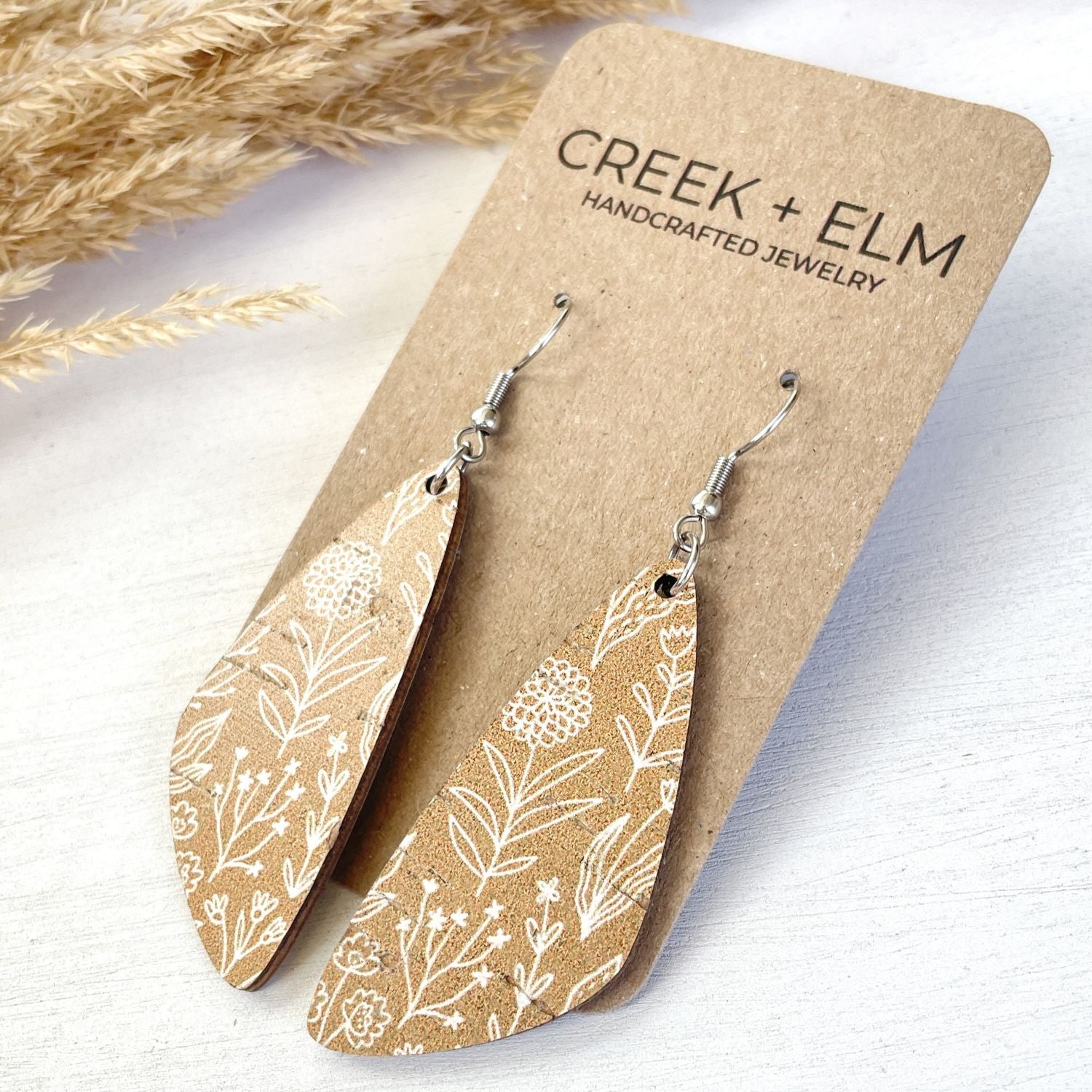 A pair of teardrop-shaped dangle earrings with a white botanical floral pattern on a tan background, displayed on a brown kraft card labeled 'CREEK + ELM HANDCRAFTED JEWELRY'.